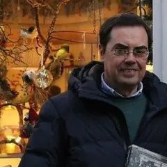 Cayo Pombo Ybarra, sonriendo, con un abrigo oscuro y un suéter verde, frente a un escaparate decorado con pájaros y adornos.