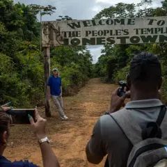 Un cartel de "Bienvenido al Templo del Pueblo Jonestown" se alza sobre un camino de tierra. Dos personas toman fotos, una de ellas posa cerca del cartel, un sombrío recordatorio de la masacre de Jonestown.