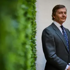 Cayetano de Borbón-Dos Sicilias, con traje gris y corbata azul, posa con una sonrisa ante una pared blanca y vegetación verde.