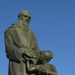 Escultura de fray Leopoldo con un niño, bajo un cielo azul. El fraile, con barba y hábito, extiende su mano hacia el pequeño.