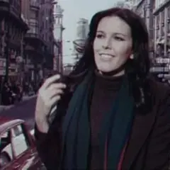 Evangelina Sobredo Galanes, con su melena oscura y bufanda, pasea por una calle urbana con edificios altos y coches antiguos.