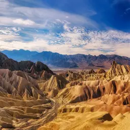 Paisaje árido y montañoso en el **Valle de la Muerte**, con formaciones rocosas erosionadas de tonos ocres y amarillos bajo un cielo azul con nubes dispersas.
