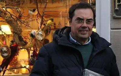 Cayo Pombo Ybarra, sonriendo, con un abrigo oscuro y un suéter verde, frente a un escaparate decorado con pájaros y adornos.
