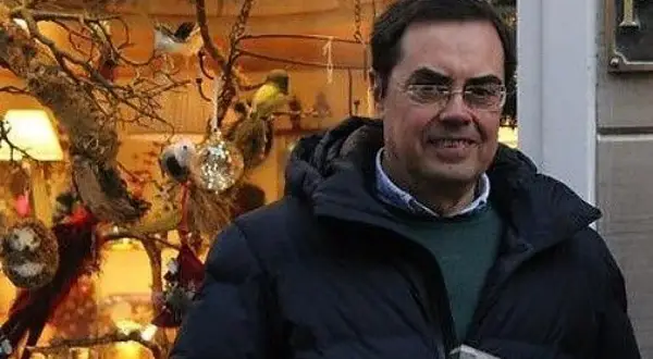 Cayo Pombo Ybarra, sonriendo, con un abrigo oscuro y un suéter verde, frente a un escaparate decorado con pájaros y adornos.