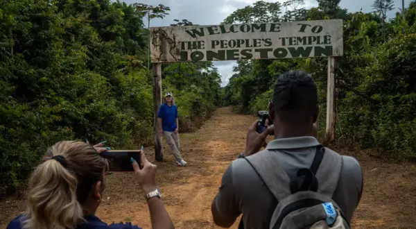 Un cartel de "Bienvenido al Templo del Pueblo Jonestown" se alza sobre un camino de tierra. Dos personas toman fotos, una de ellas posa cerca del cartel, un sombrío recordatorio de la masacre de Jonestown.