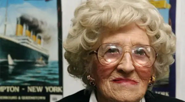 Una anciana con gafas grandes sonríe. Detrás de ella, carteles del Titanic, evocando a los supervivientes del trágico viaje.