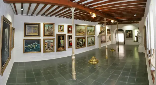 Galería de arte con cuadros y esculturas, techos de madera y suelo de baldosas.