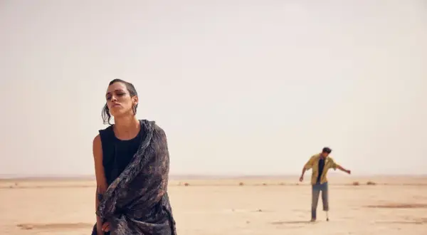 Mujer con pañuelo en desierto, hombre con muleta al fondo. El sirat, significado de un camino difícil.
