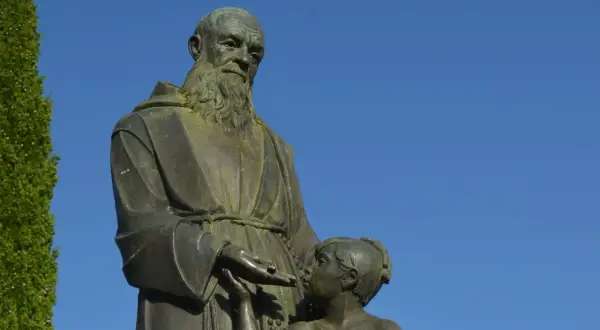 Escultura de fray Leopoldo con un niño, bajo un cielo azul. El fraile, con barba y hábito, extiende su mano hacia el pequeño.