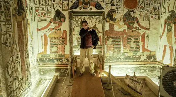 Un hombre posa en la tumba de Nefertari, rodeado de vibrantes jeroglíficos y figuras divinas que narran su viaje al más allá.