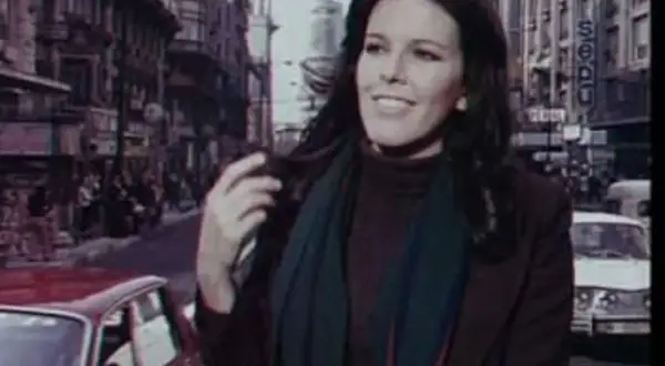 Evangelina Sobredo Galanes, con su melena oscura y bufanda, pasea por una calle urbana con edificios altos y coches antiguos.