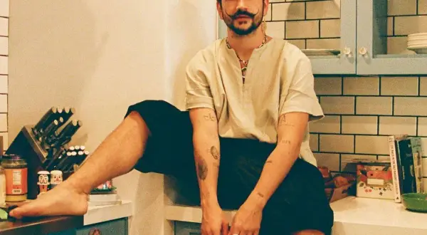 Camilo Alonso Vega, con su característico bigote, posa relajado en una cocina. Sus tatuajes y anillos añaden un toque personal a su estilo.