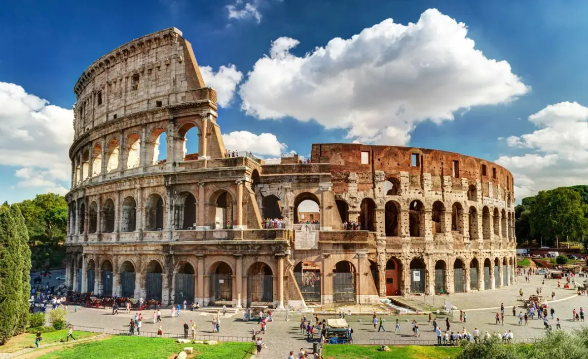 El **Coliseo de Roma**, imponente anfiteatro romano, recibe a turistas bajo un cielo azul con nubes.