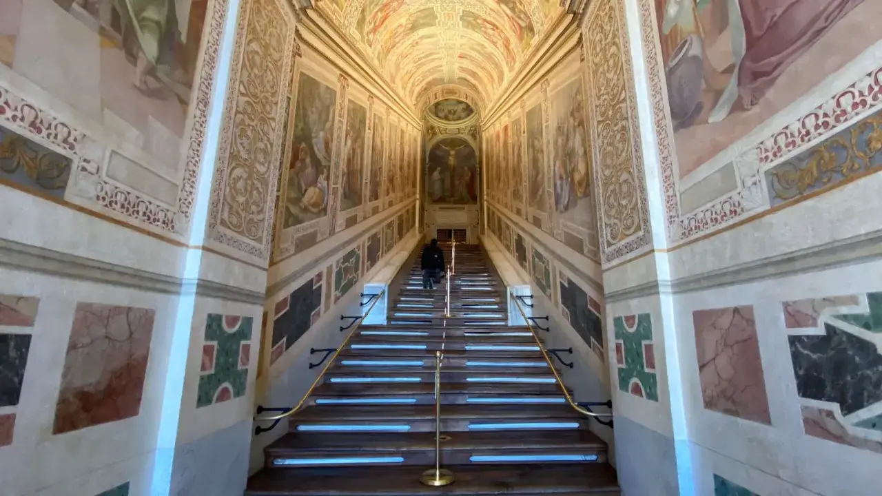 Una persona sube la escalera santa en Roma, rodeada de frescos y m&aacute;rmoles.