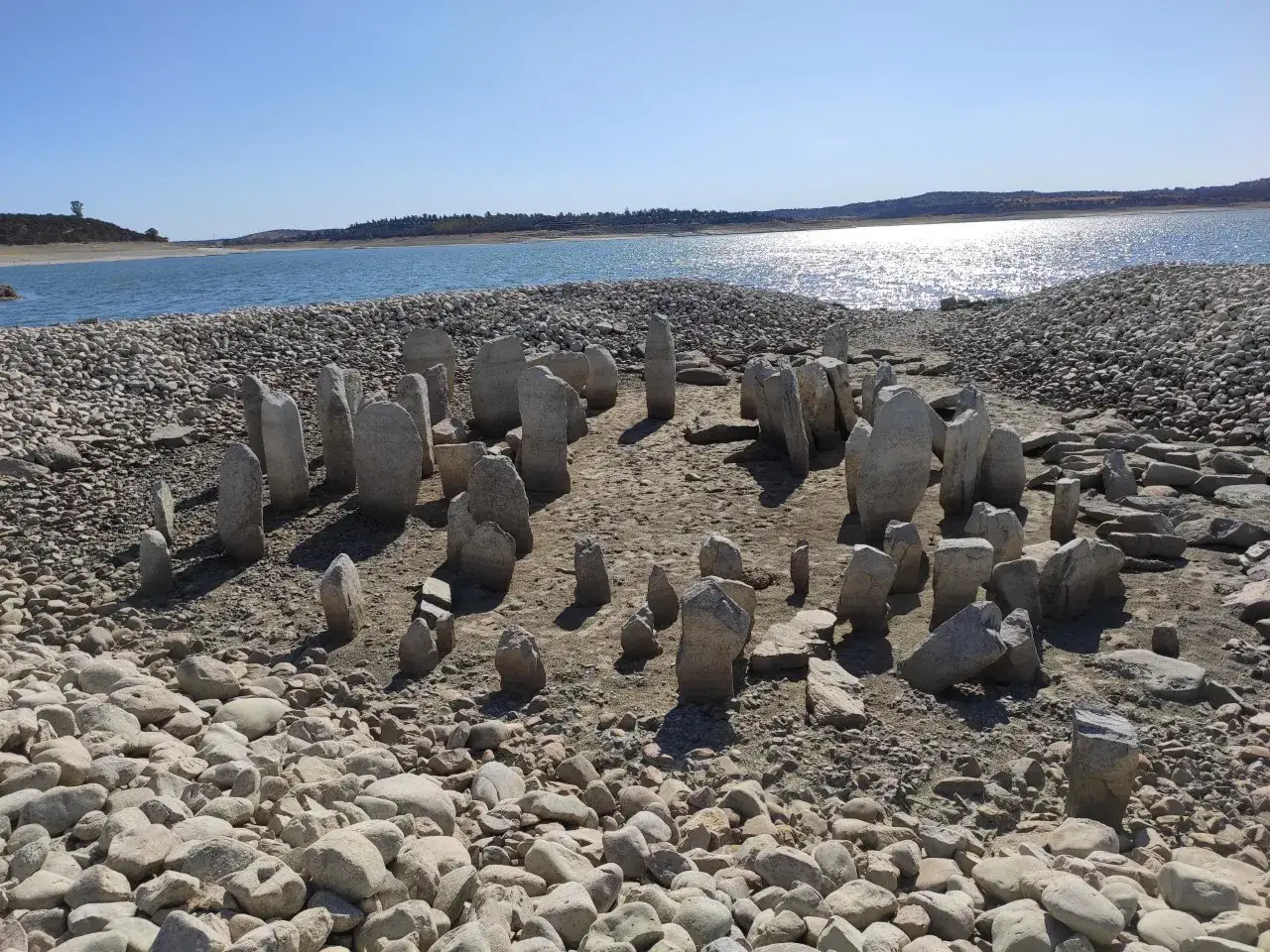 El dolmen de Guadalperal emerge de las aguas bajas, un c&iacute;rculo de piedras ancestrales bajo un cielo azul.