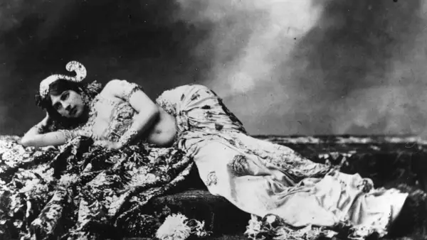Mata Hari, bailarina exótica, yace recostada sobre telas estampadas, con un tocado adornado y mirada cautivadora.