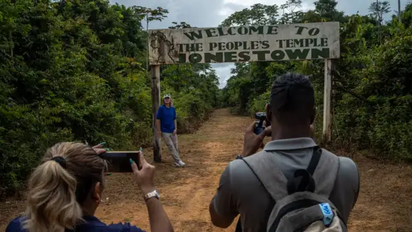 Un cartel de "Bienvenido al Templo del Pueblo Jonestown" se alza sobre un camino de tierra. Dos personas toman fotos, una de ellas posa cerca del cartel, un sombrío recordatorio de la masacre de Jonestown.