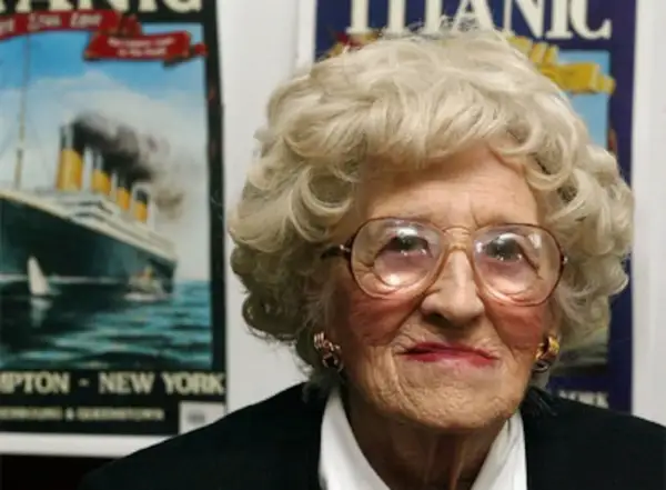 Una anciana con gafas grandes sonríe. Detrás de ella, carteles del Titanic, evocando a los supervivientes del trágico viaje.
