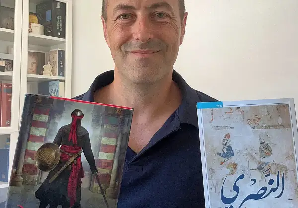 Juan de Granada, sonriente, sostiene dos libros. Uno muestra un guerrero con armadura y el otro, una escena histórica.