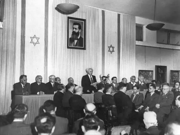 Hombres en traje asisten a un evento histórico, la creacion del estado de Israel, con la bandera israelí y un retrato de Theodor Herzl.