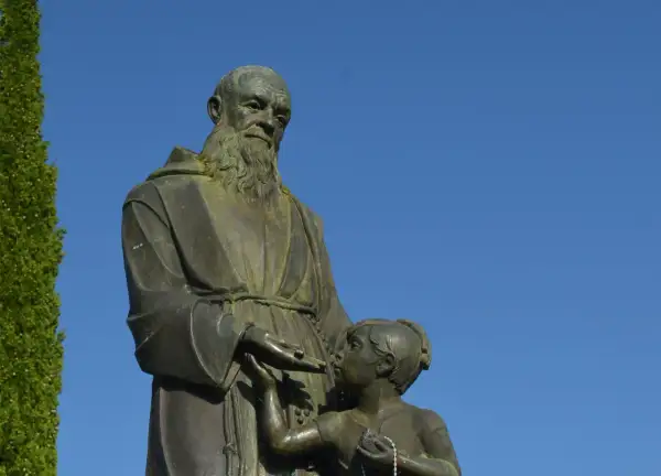 Escultura de fray Leopoldo con un niño, bajo un cielo azul. El fraile, con barba y hábito, extiende su mano hacia el pequeño.
