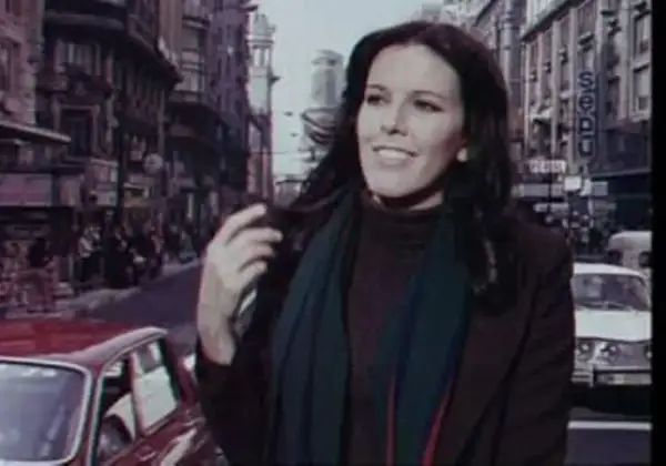 Evangelina Sobredo Galanes, con su melena oscura y bufanda, pasea por una calle urbana con edificios altos y coches antiguos.