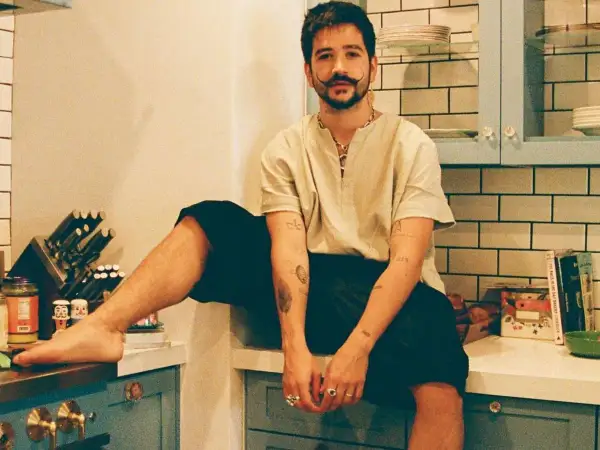 Camilo Alonso Vega, con su característico bigote, posa relajado en una cocina. Sus tatuajes y anillos añaden un toque personal a su estilo.